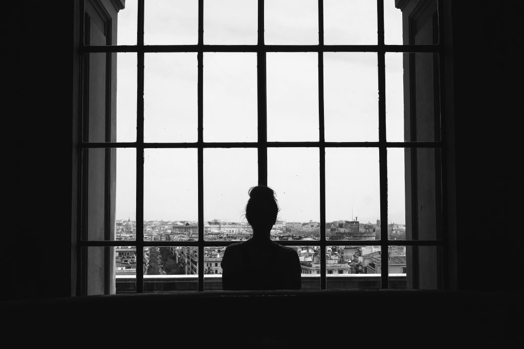 photo en noir et blanc d'une femme seule debout devant les fenêtres, regardant les bâtiments.