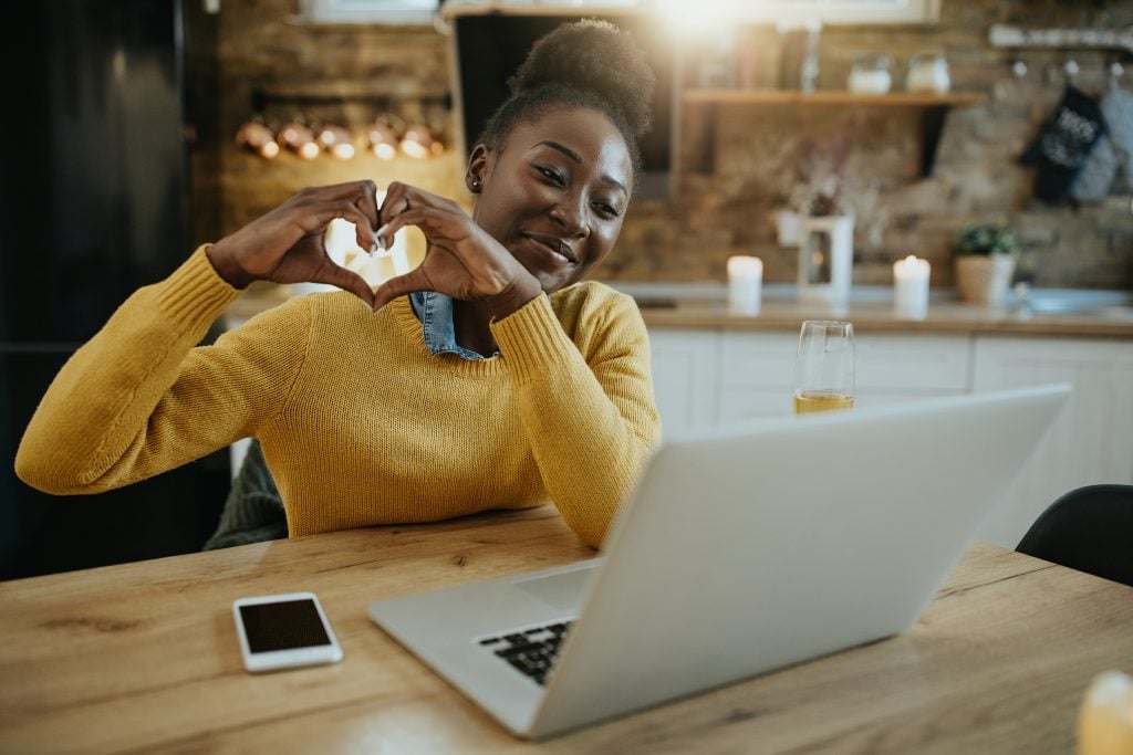 Femme afro-américaine heureuse utilisant un ordinateur lors d'un rendez-vous en ligne et montrant une forme de cœur à quelqu'un.